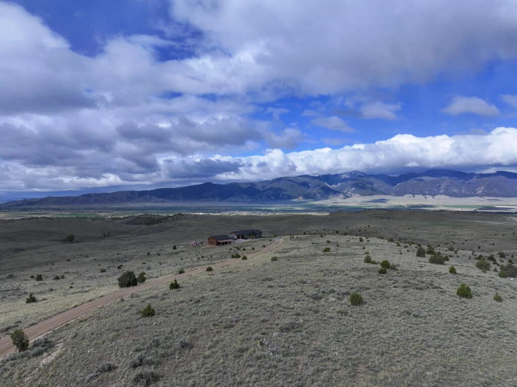 A rural landscape features a dirt road—380 Mill Gulch Road—leading to a cluster of houses amid rolling grassy hills, with scattered shrubs and distant mountains under a partly cloudy sky.