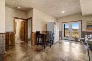A rustic kitchen at 380 Mill Gulch Road with wooden paneling and concrete floors features a small table, refrigerator, and double glass doors opening to a scenic mountain view. A bathroom is visible to the left.