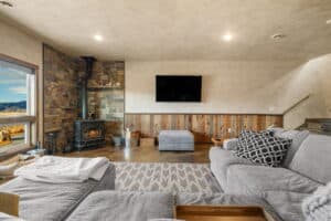 A cozy living room at 380 Mill Gulch Road features a gray sectional sofa, patterned cushions, a wood-burning stove against a stone wall, a flat-screen TV, and a large window framing scenic mountain views.