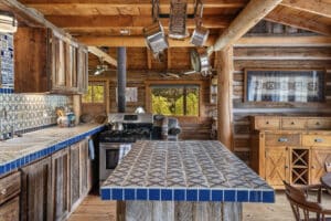 Rustic log cabin kitchen at a Montana retreat, with patterned blue and white tiled countertops, wooden cabinets, pots hanging above the island, a stove, and a window overlooking Tom Miner Creek’s lush greenery. Warm wood tones throughout.