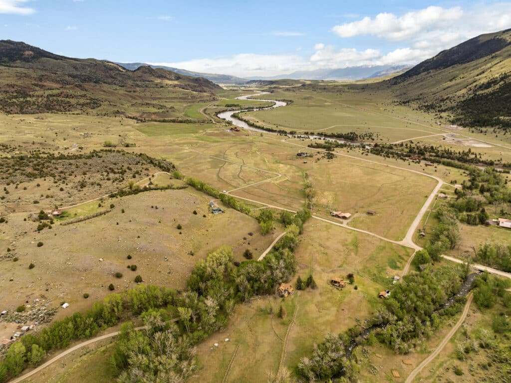 Aerial view of Tom Miner Creek winding through a wide Montana valley with scattered trees, grassy fields, and a few houses. Rolling hills and mountains surround this peaceful retreat under a partly cloudy sky.