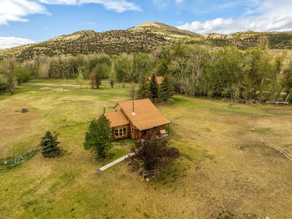 A small wooden cabin with a red roof sits in an open grassy field near Tom Miner Creek, surrounded by trees, hills, and mountains under a partly cloudy blue sky.