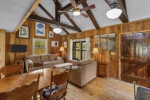 Cozy rustic living room retreat with wood-paneled walls, exposed beams, a ceiling fan, two beige sofas, a wooden dining table with chairs, landscape art, lamps, and windows filling the Montana space with natural light.