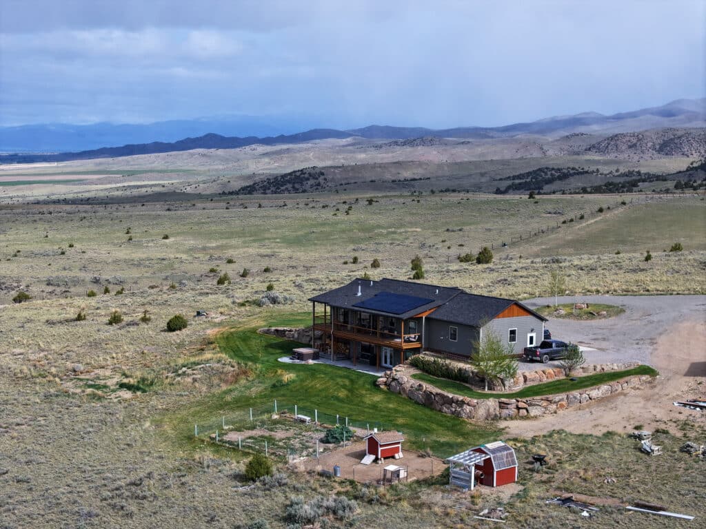 Nestled at 380 Mill Gulch Road, this farmhouse with a wraparound porch sits in a vast landscape with mountain views. Enjoy green lawns, a garden, small red outbuildings, and dry grassy fields under a cloudy sky.