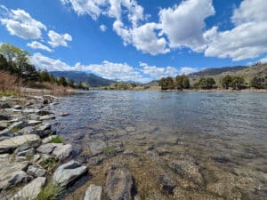 Clear river with visible stones at the shore, surrounded by grassy banks and trees, with distant hills and mountains under a partly cloudy blue sky near Tom Miner Creek Retreat.