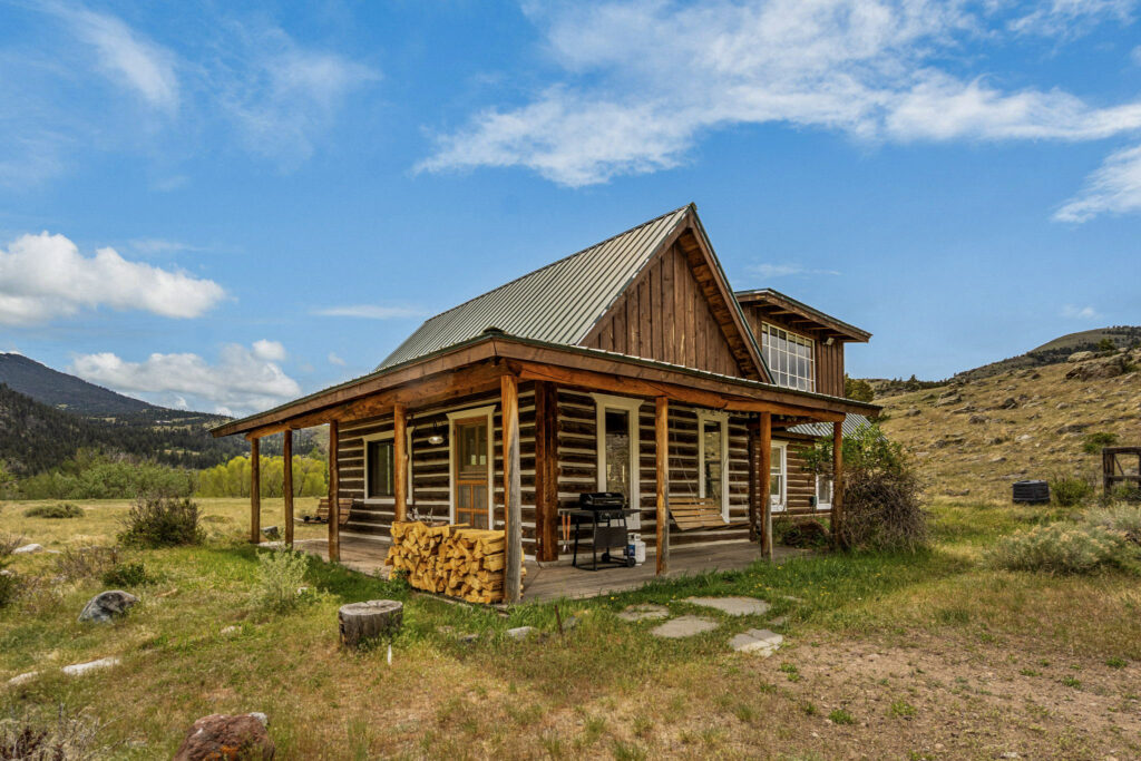 A rustic log cabin with a metal roof sits in a grassy, open landscape at Tom Miner Creek Retreat. There’s a covered porch with firewood stacked beside it, and mountains and blue sky with clouds in the background.