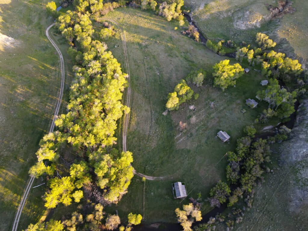 Aerial view of a rural landscape with winding dirt roads, scattered small cabins, green fields, and clusters of trees bordered by a narrow stream—ideal as a hunting property or ranch for sale. Sunlight casts long shadows across the scene.