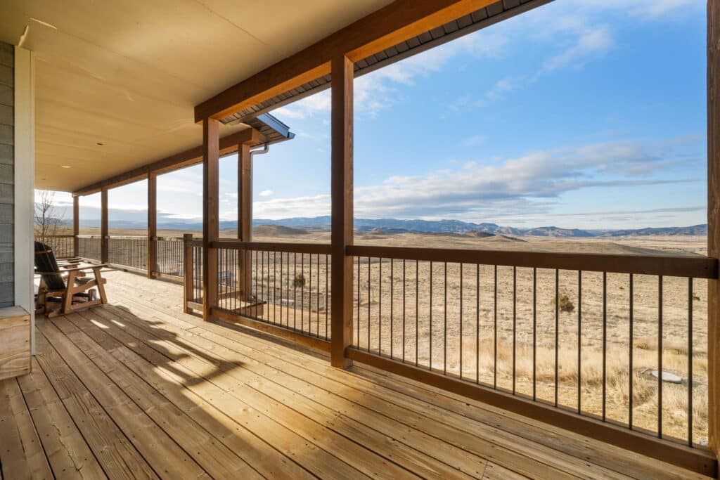 The spacious wooden porch at 380 Mill Gulch Road, with its railing, overlooks a wide open field and distant mountains. Two chairs invite relaxation beneath a blue sky dotted with clouds, perfect for enjoying the tranquil scenic view.