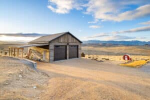 A wooden garage with two brown doors and a covered side area stands on a dirt lot at 380 Mill Gulch Road, set in a dry, open landscape. In the distance, there’s a small red shed and rolling hills beneath a blue sky with scattered clouds.