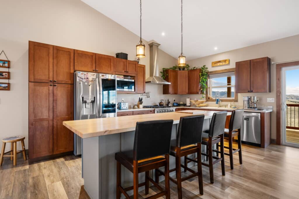 Modern kitchen at 380 Mill Gulch Road with wooden cabinets, stainless steel appliances, a spacious island with a light wood countertop, four dark chairs, pendant lights, and a window showcasing a scenic view in the background.