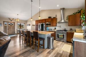 Modern kitchen at 380 Mill Gulch Road with wooden cabinets, stainless steel appliances, an island with barstools, pendant lights, and a cozy dining area. Open layout features wood flooring, large windows for natural light, and a wall-mounted bull skull decor.