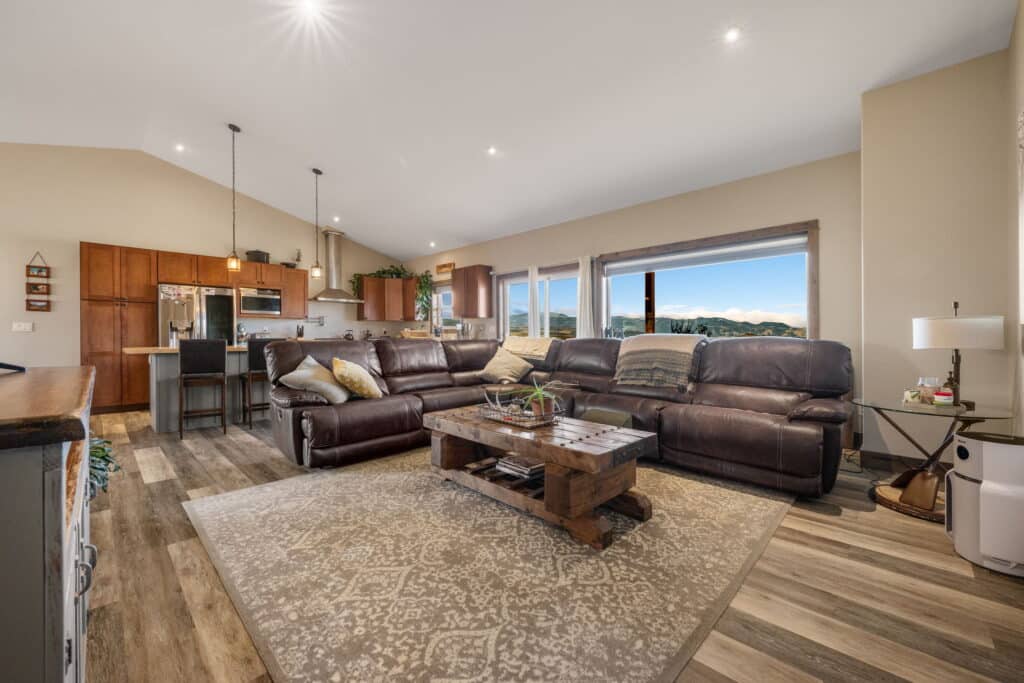 Spacious open-concept living room and kitchen at 380 Mill Gulch Road, featuring a large brown sectional sofa, rustic coffee table, modern kitchen appliances, bar stools, and expansive windows with scenic mountain views.