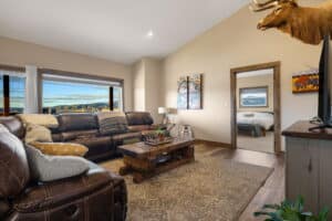 A cozy living room at 380 Mill Gulch Road features brown leather sofas, a rustic wooden coffee table, and animal-themed decor. Large windows showcase scenic mountain views, with an adjoining bedroom visible through an open doorway.