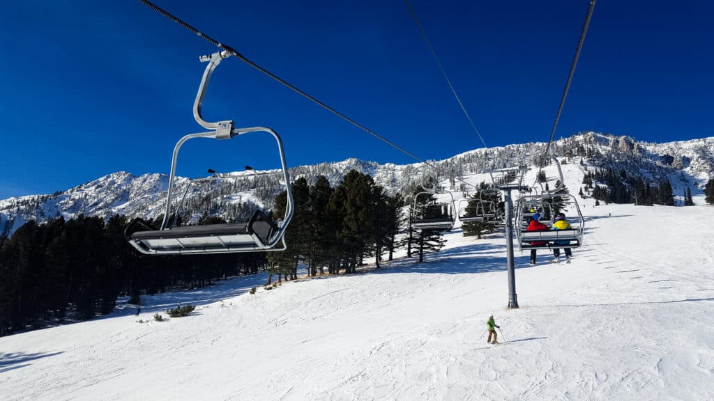 A ski lift glides over a snowy mountain slope in Bridger Canyon, with pine trees and skiers below, under a clear blue sky and rugged mountains—a perfect winter scene near Spring Creek Estate.