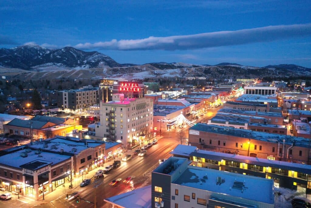 A winter aerial view of a small city at dusk features illuminated streets, snow-covered rooftops, and the prominent Hotel Baxter sign. The mountains near Bridger Canyon rise in the background under a partly cloudy sky.