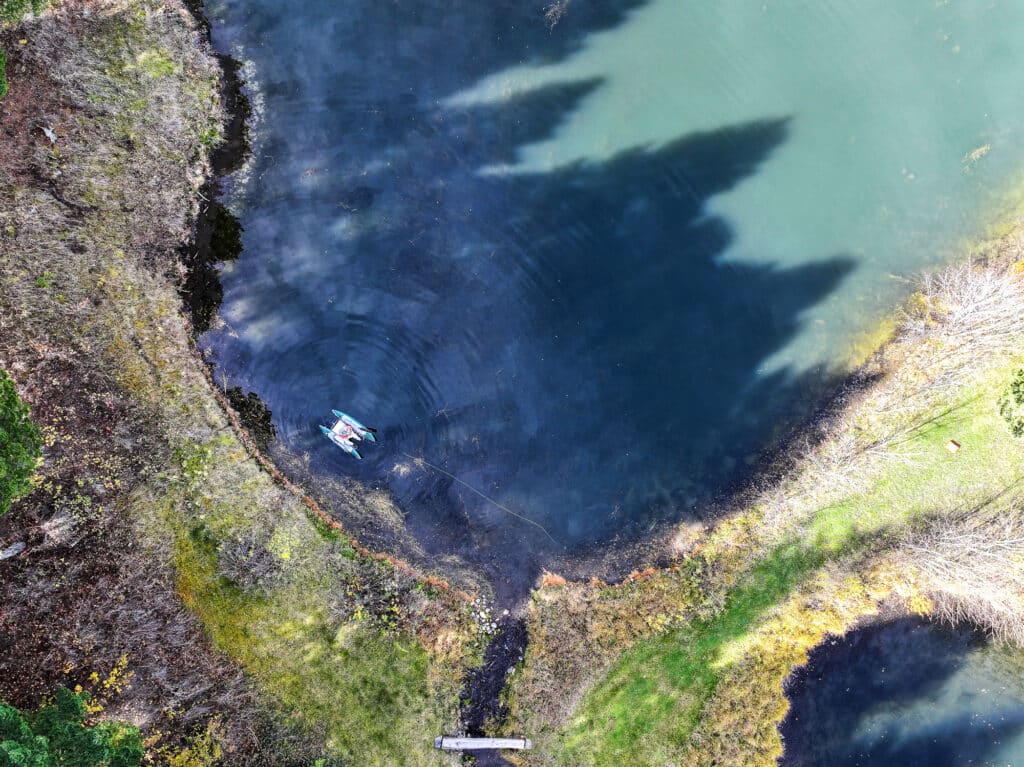 Aerial view of a small boat with a person near the edge of Spring Creek, surrounded by grass and trees in Bridger Canyon. Tree shadows stretch over the water, and an estate dock is visible at the bottom of the image.
