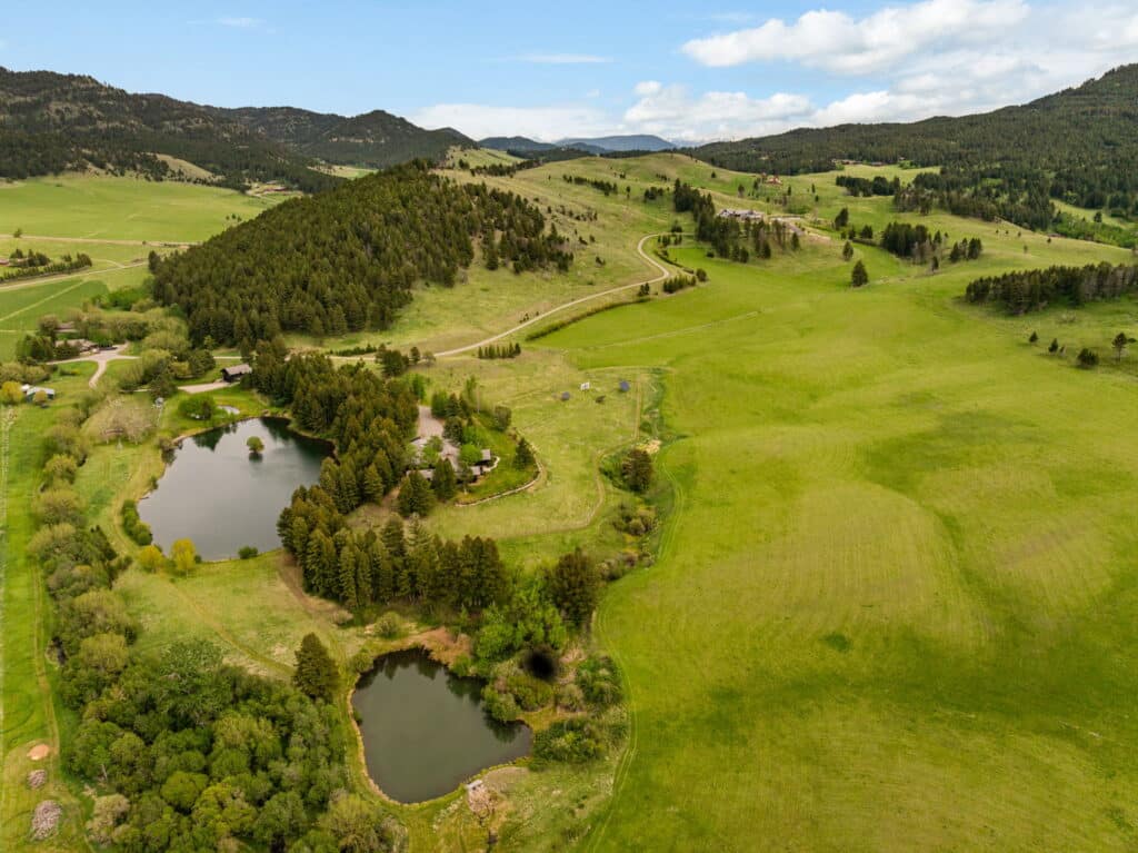 Aerial view of a lush green landscape with rolling hills, scattered trees, two small ponds, and a winding road stretching into the distance under a partly cloudy sky.