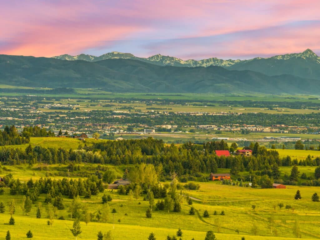 Rolling green hills dotted with trees and houses in the foreground, a town in the middle distance, and the majestic Five Ranges with snow-capped peaks under a pink and blue sunset sky in the background.