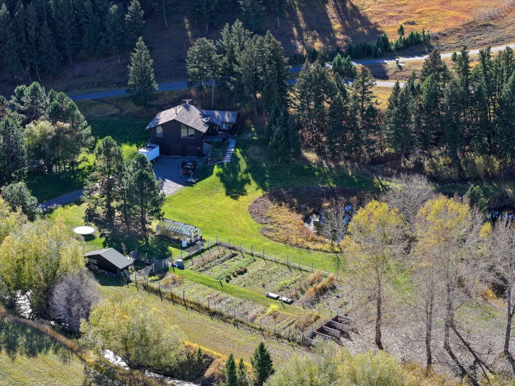 Aerial view of a rural property in Bridger Canyon with a house, driveway, garden with raised beds, greenhouse, trees, and a road in the background surrounded by greenery and forest—ideal Bridger Canyon real estate.