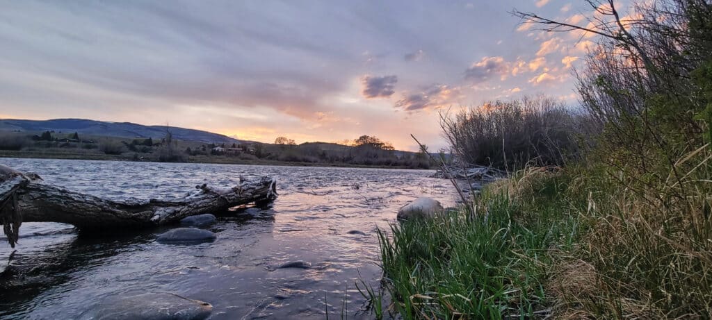 A peaceful river flows past rocks and a fallen tree at sunset, with grass and bushes lining the bank. In the distance, hills near Sypes Canyon rise under a colorful, partly cloudy sky.