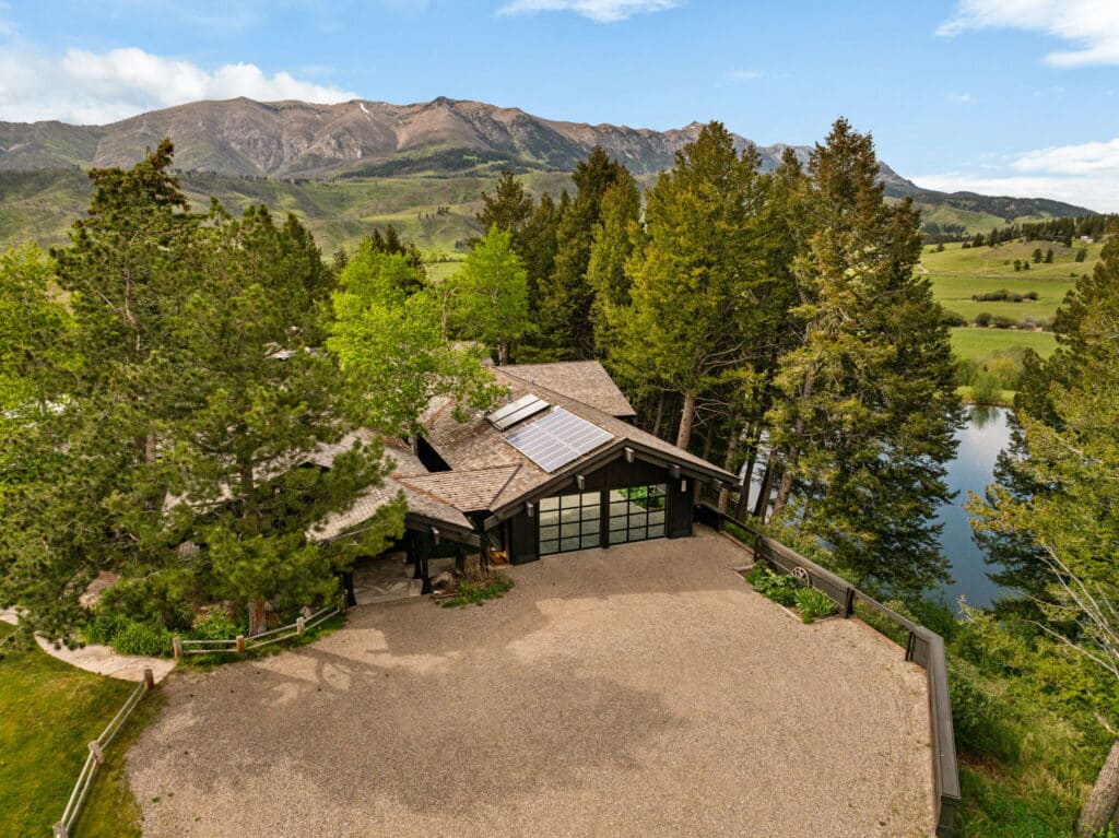 A modern house with large glass windows sits surrounded by tall trees on the Spring Creek Estate, overlooking a pond and green hills, with mountains in the background under a blue sky.