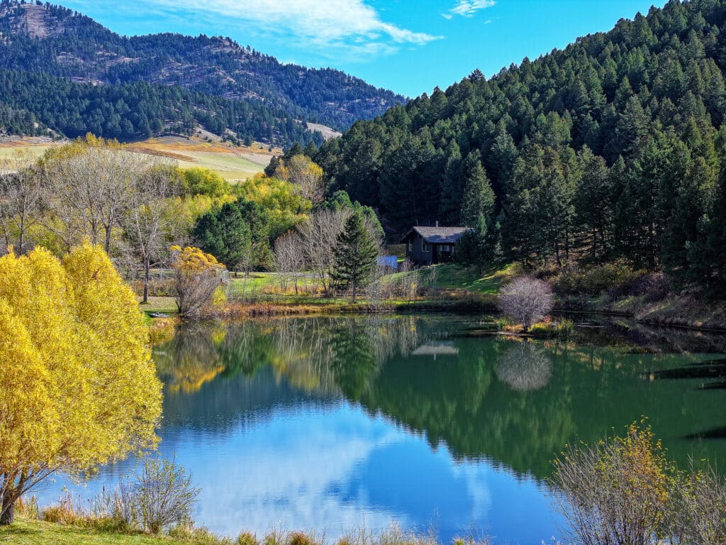 A serene lake reflects surrounding trees, some with autumn leaves, and a mountain range under a bright blue sky. A small house is nestled among the trees near the water’s edge on this peaceful Bridger Canyon estate.