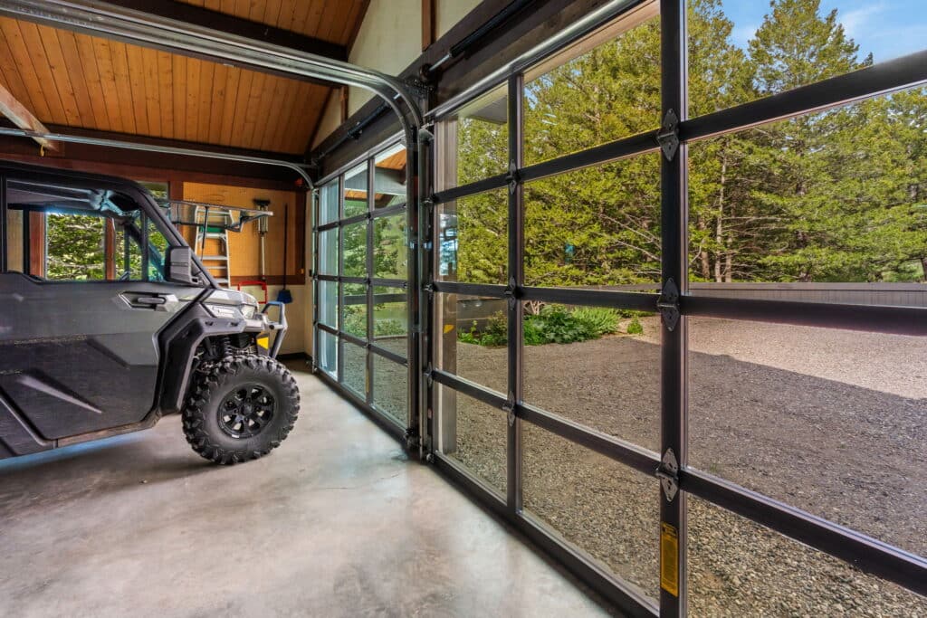 A garage with glass-paneled doors opens to a gravel driveway, surrounded by trees in Bridger Canyon. An off-road vehicle with large tires is parked inside on a concrete floor.