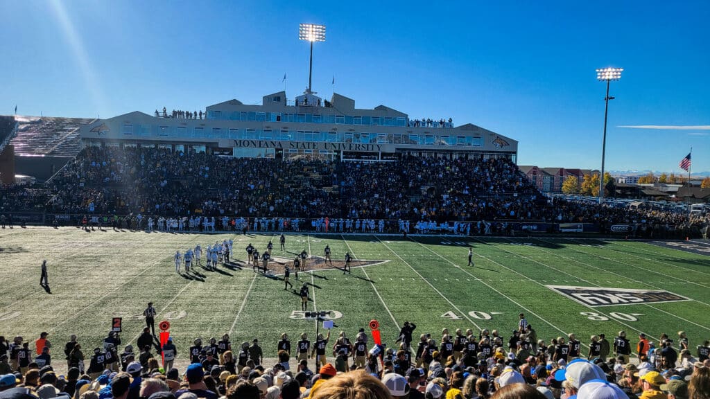 A college football game at Montana State University shows players on the field and a crowd of fans in the stadium under a clear blue sky, with the university building and Bridger Canyon visible in the background.