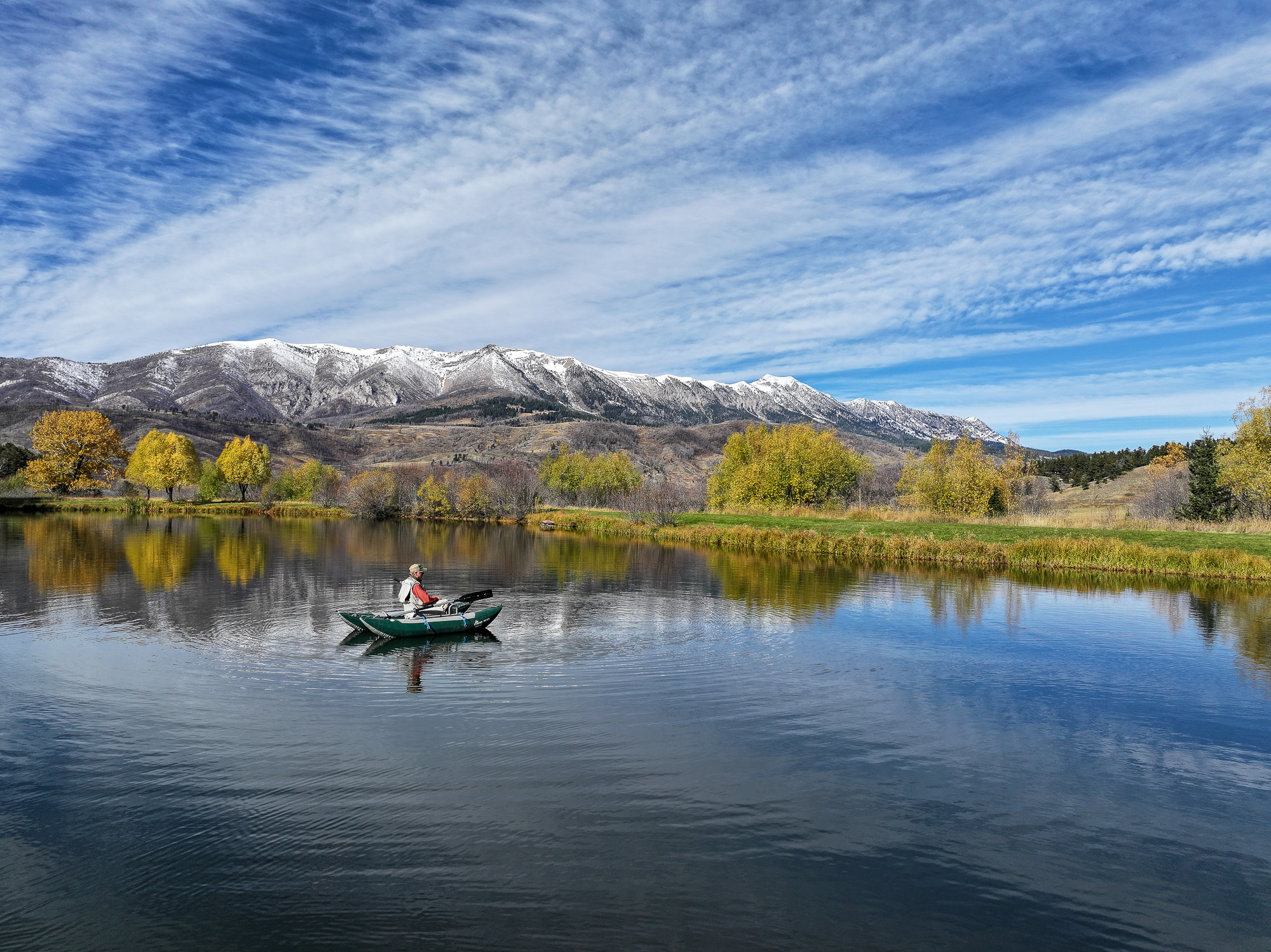 A person in a small green boat rows on a calm lake near Spring Creek, surrounded by autumn trees, with snow-capped mountains and a blue sky with wispy clouds in the background.