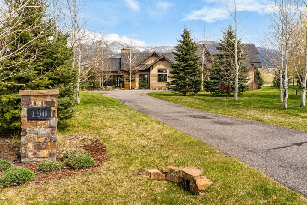 A stone marker with the number 190 stands by a paved driveway leading to a large house on Jeana Lei Court, surrounded by green grass, trees, and mountain views—an inviting real estate opportunity under a clear blue sky.