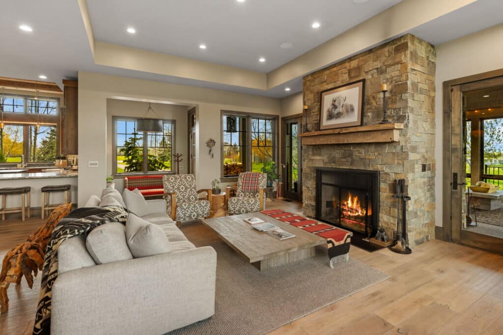 A cozy living room at 190 Jeana Lei Court features a stone fireplace, beige sofa, armchairs, wooden coffee table, and large windows with views of trees. Warm lighting and rustic decor make this real estate opportunity inviting and charming.