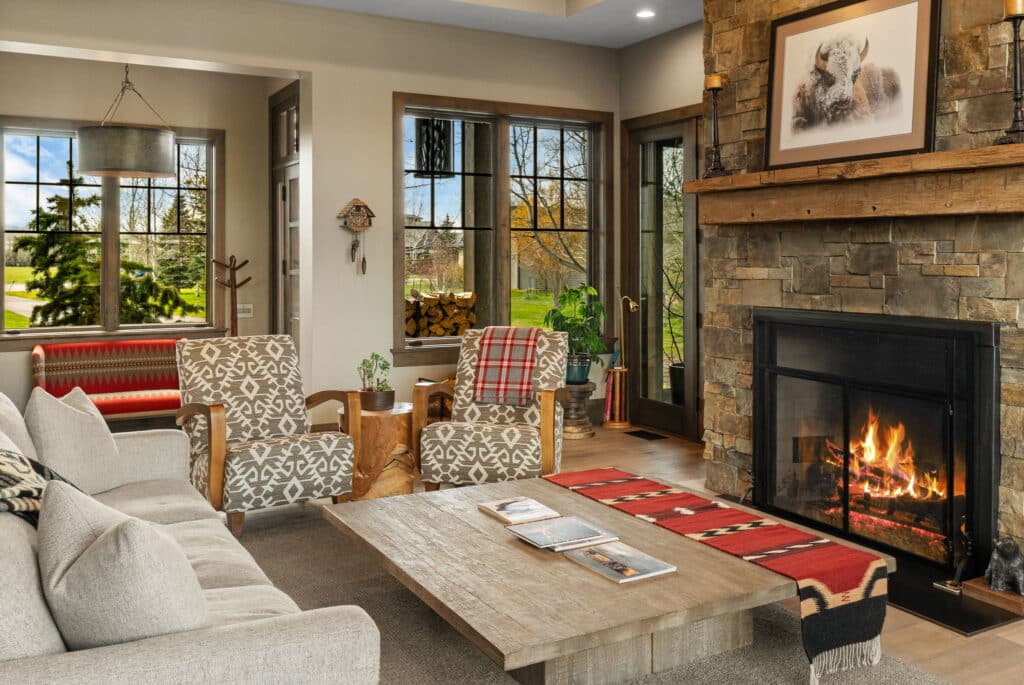 A cozy living room in Jeana Lei Court features a stone fireplace with fire burning, wooden coffee table, patterned armchairs, beige sofa, red-striped accents, large windows overlooking greenery, and a photo of a bear above the mantel.