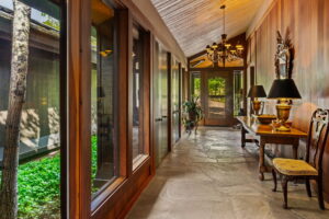 A hallway with stone flooring and wood-paneled walls, this Estate features large windows overlooking a garden, a decorative console table with lamps and a mirror, chairs, plants, and a chandelier, all leading to a glass door.