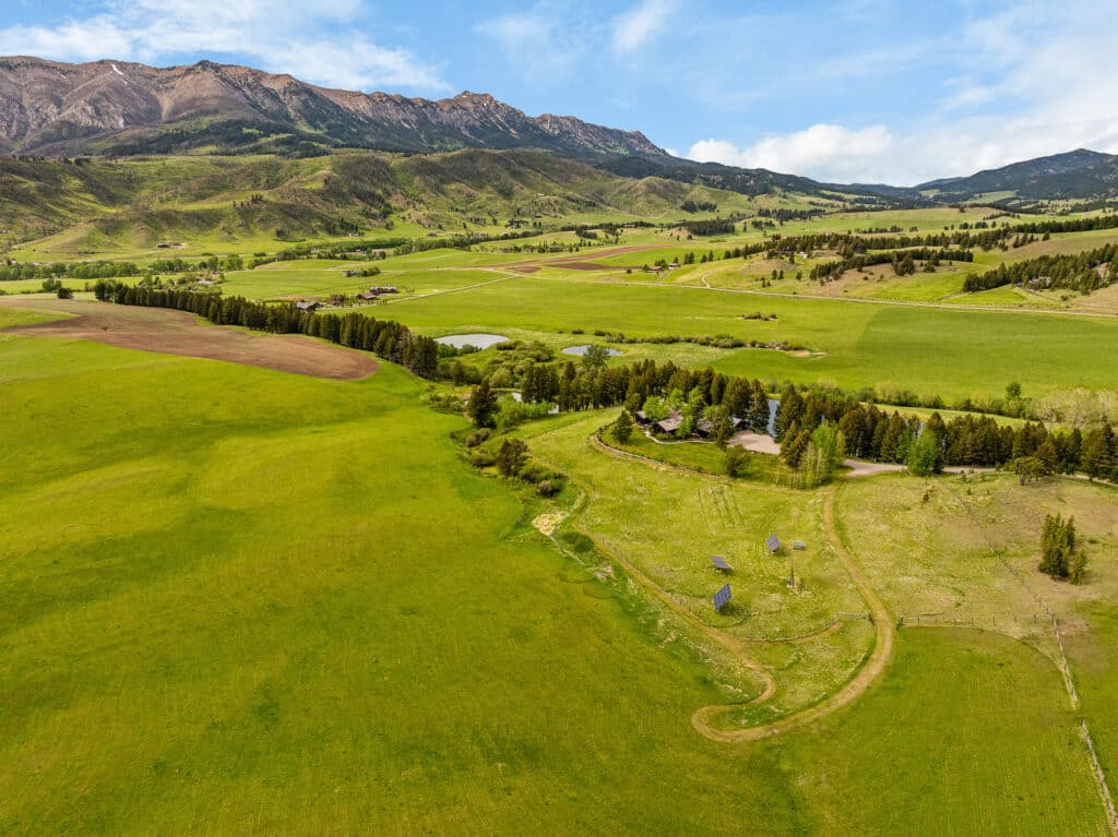 Aerial view of a lush green valley in Bridger Canyon with scattered trees, ponds, and a few estate homes, surrounded by rolling hills and mountains under a blue sky with clouds.