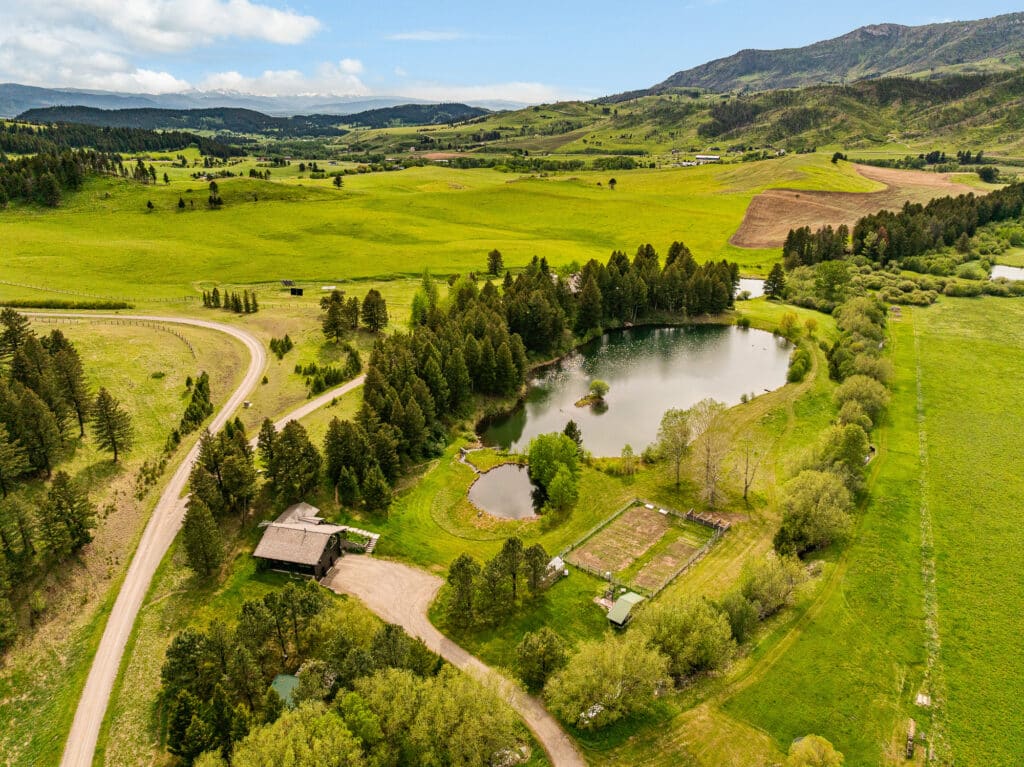Aerial view of a rural estate in Bridger Canyon with a house, two ponds, trees, garden area, fields, and a winding dirt road amid rolling green hills and distant mountains under a partly cloudy sky.