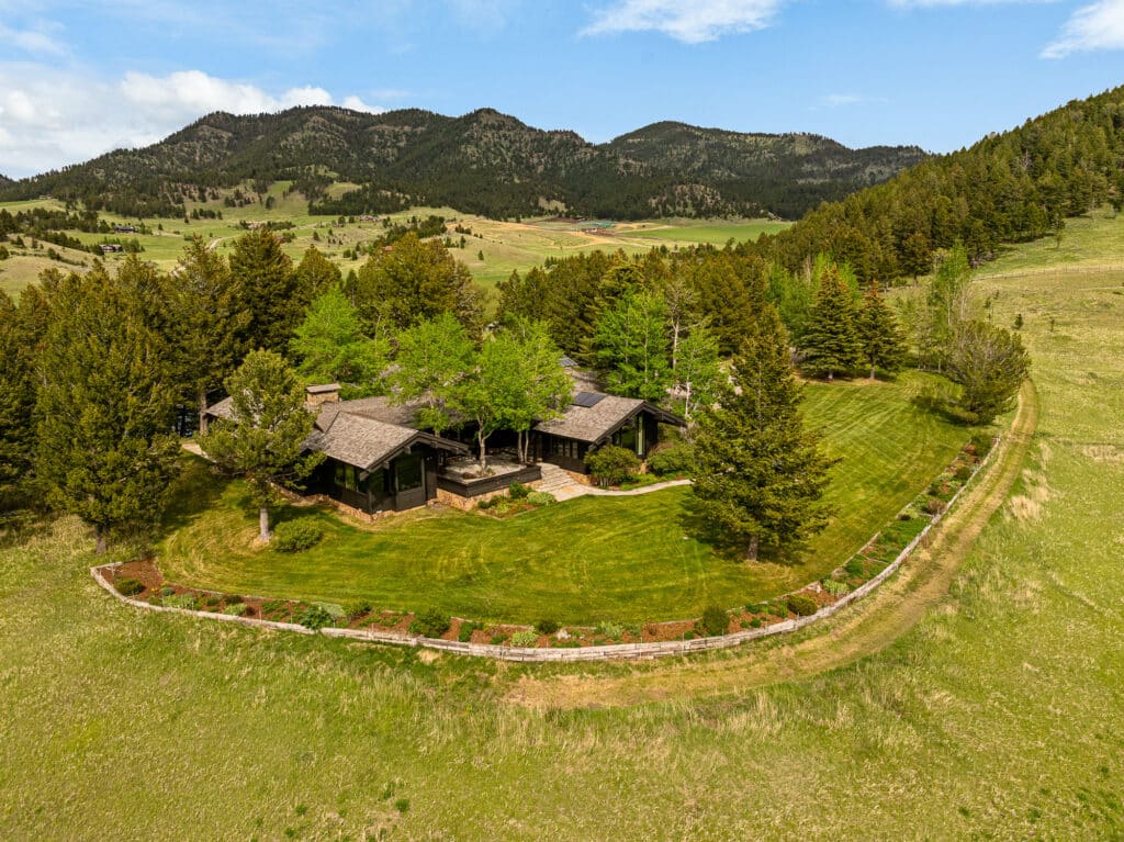 A rustic estate surrounded by lush trees and manicured lawns sits in a fenced area amid grassy hills and mountains of Bridger Canyon under a partly cloudy sky.