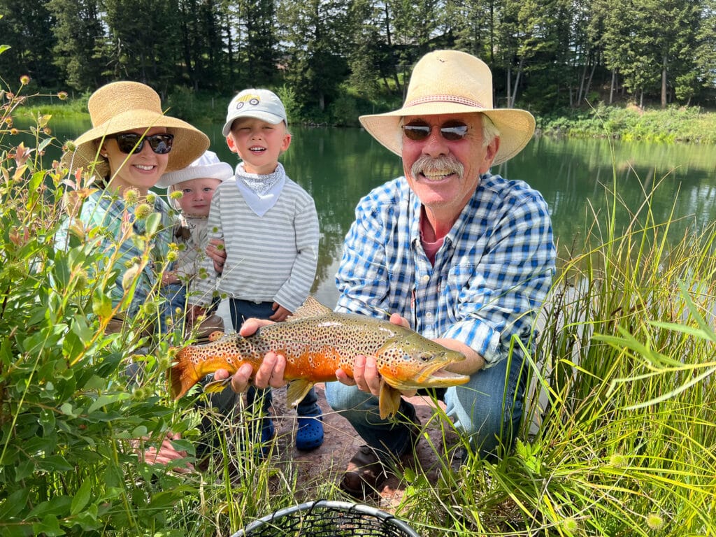 A smiling man in a straw hat and plaid shirt kneels by a pond at Spring Creek, holding a large brown trout. A woman and two young children, also in sun hats, stand beside him among green grass and wildflowers.