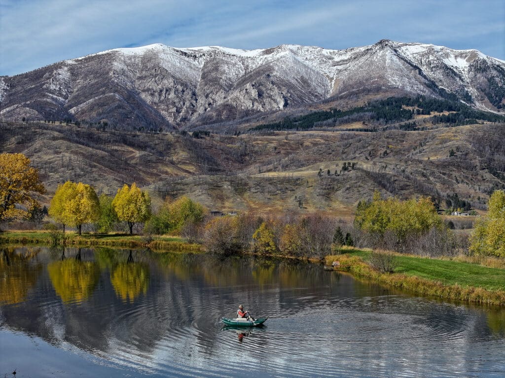 A person rows a small boat on a calm lake in Bridger Canyon, surrounded by autumn trees, with snow-capped mountains and a partly cloudy sky in the background. The colorful foliage and mountains are reflected in the water.