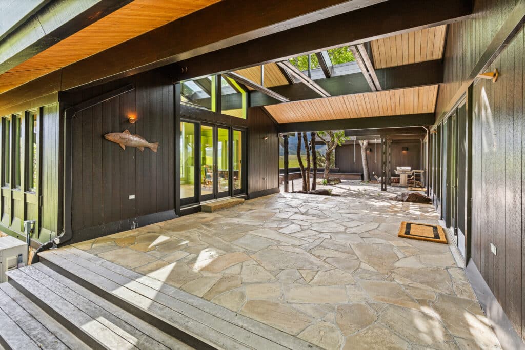 Spacious covered patio with stone flooring, dark wood walls, large windows, and skylights. A fish wall decoration and doormat are visible. Enjoy views of greenery through the glass in this Bridger Canyon estate retreat.