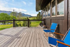A wooden deck with two blue chairs overlooks a lush green yard, trees, and distant Bridger Canyon mountains under a blue sky with scattered clouds. Large windows line the side of this inviting estate adjacent to the deck.