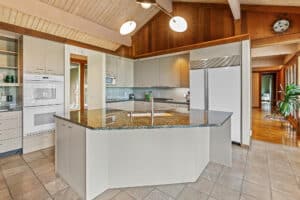 Spacious Bridger Canyon kitchen with a large granite island, white cabinets, built-in oven and microwave, double-door fridge, tiled floor, and wood-paneled vaulted ceiling with exposed beams and modern lighting.