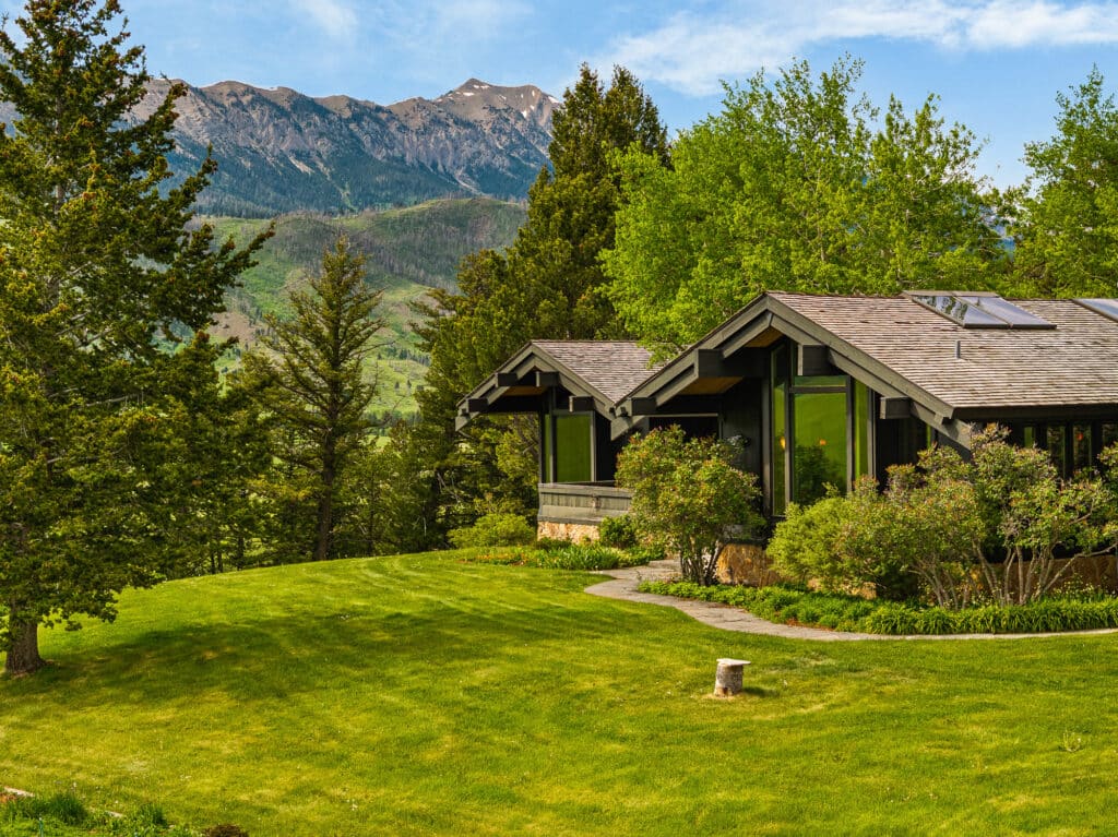 A modern house with large windows sits on a well-manicured green lawn in the Spring Creek Estate, surrounded by trees and shrubs, with mountains visible in the background under a partly cloudy Bridger Canyon sky.