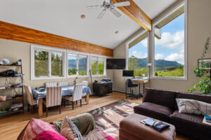 A bright living-dining area in Bridger Canyon features large windows with mountain views, a dining table with checkered cloth, a dark sectional sofa, a rug, a desk with computer, and a ceiling fan under the sloped wooden beam ceiling.
