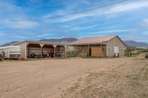 A rural scene at 868 Middle Road with a beige metal barn and open-sided shed storing farm equipment, surrounded by dirt ground and hay bales, set against mountains and a clear blue sky.