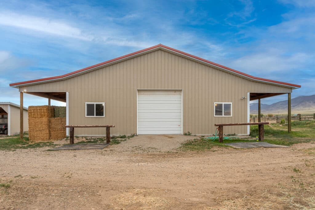 A beige metal barn at 868 Middle Road features a white roll-up door, two side shelters, hay bales stacked on the left, and open land with mountains in the background under a partly cloudy sky.