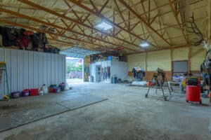 A spacious, well-lit barn interior at 868 Middle Road features an open roll-up door, exposed beams, saddle racks, buckets, and various tools and supplies neatly organized along the walls.