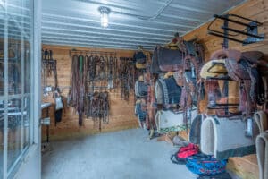 A tack room at 868 Middle Road features saddles, bridles, and horse gear neatly organized on wooden walls and racks. The metal ceiling, concrete floor, and fluorescent light complete the space, while a few saddle pads and bags rest on the floor.