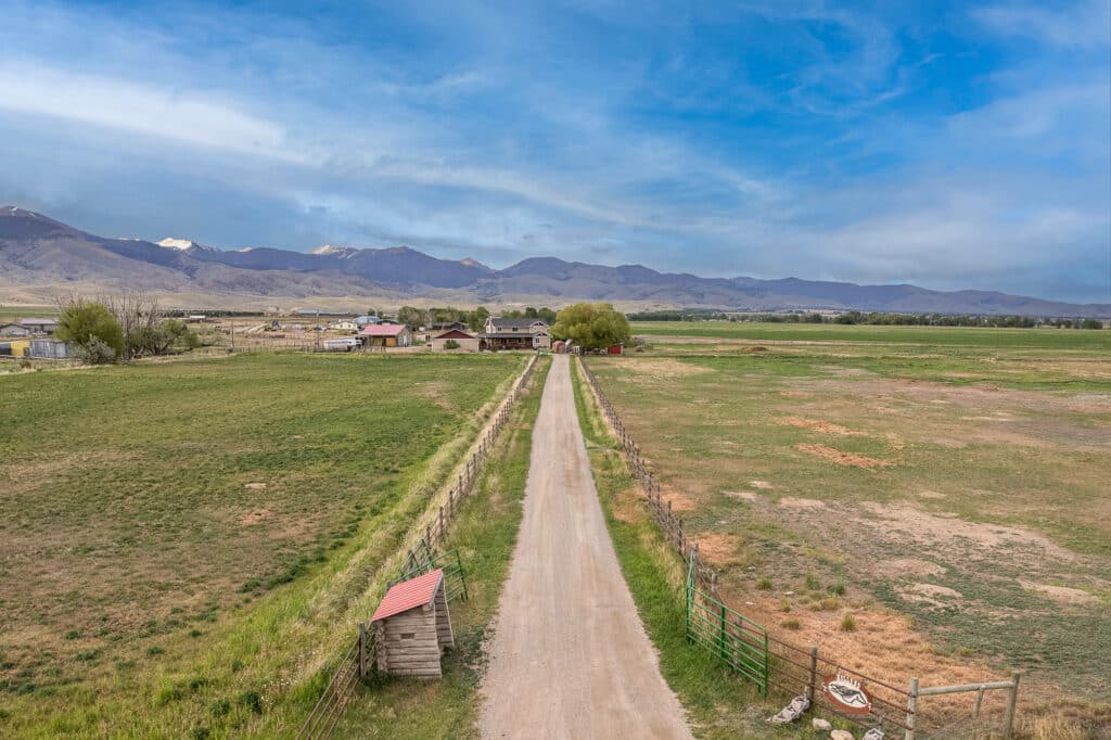 A long dirt road, known as 868 Middle Road, leads to a farmhouse surrounded by green fields and fenced pastures, with distant mountains and a blue sky in the background.