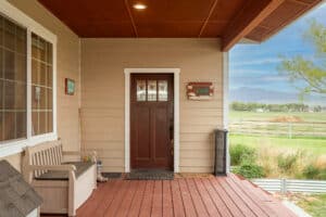 A cozy porch at 868 Middle Road, with a brown wooden door, a bench, and welcome signs, overlooks a scenic rural landscape with green fields, trees, and distant mountains under a blue sky.