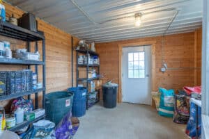 A small storage room at 868 Middle Road with wooden walls, metal ceiling, plastic bins, shelves holding various containers, bags of animal feed, and a door with a window letting in daylight.